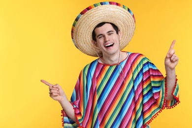 Young man in Mexican sombrero hat and poncho pointing at something on yellow background