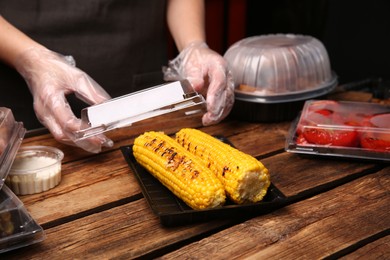 Photo of Waiter in gloves closing container with grilled corn cobs at wooden table, closeup. Food delivery service