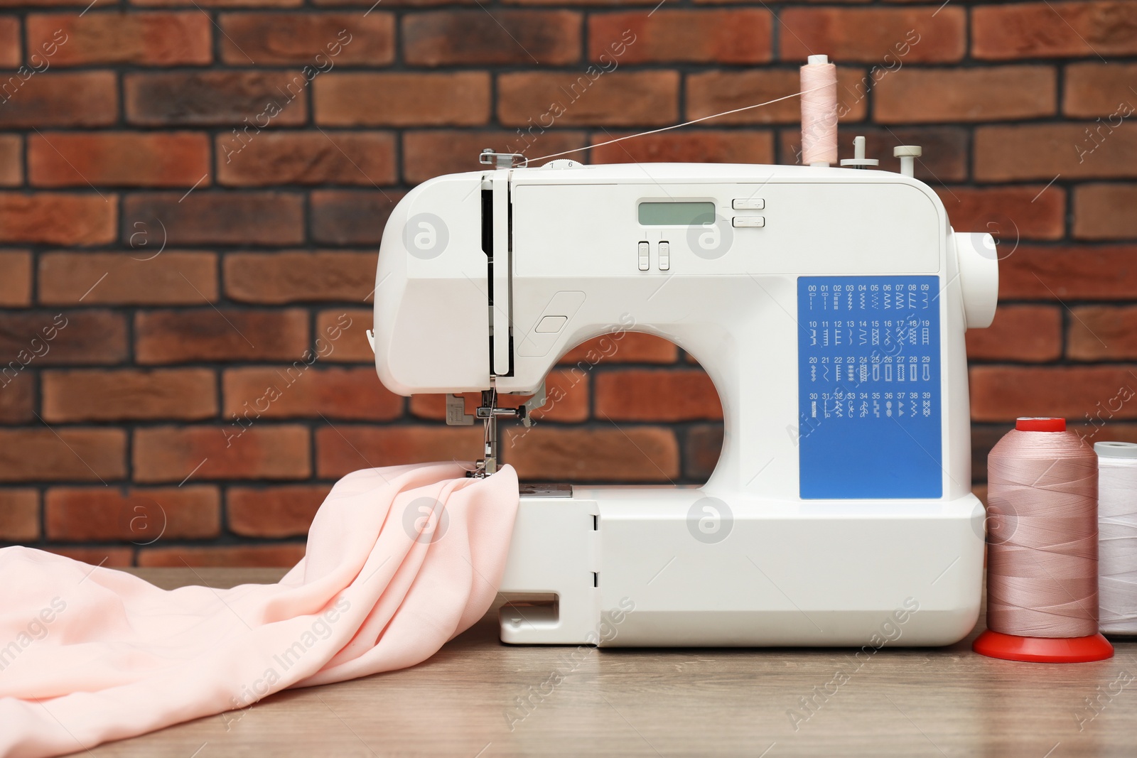 Photo of Sewing machine with fabric and spools of threads on wooden table against brick wall