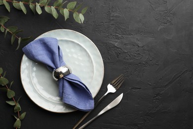 Stylish setting with cutlery, eucalyptus branches, napkin and plate on dark textured table, flat lay. Space for text