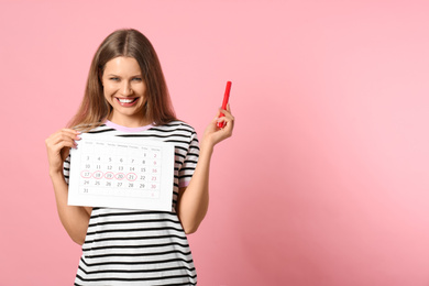Photo of Young woman holding calendar with marked menstrual cycle days on pink background. Space for text
