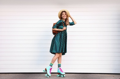 Happy stylish young woman with vintage roller skates, hat and backpack near white garage door on street