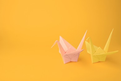 Origami art. Beautiful pale pink and light yellow paper cranes on orange background, space for text