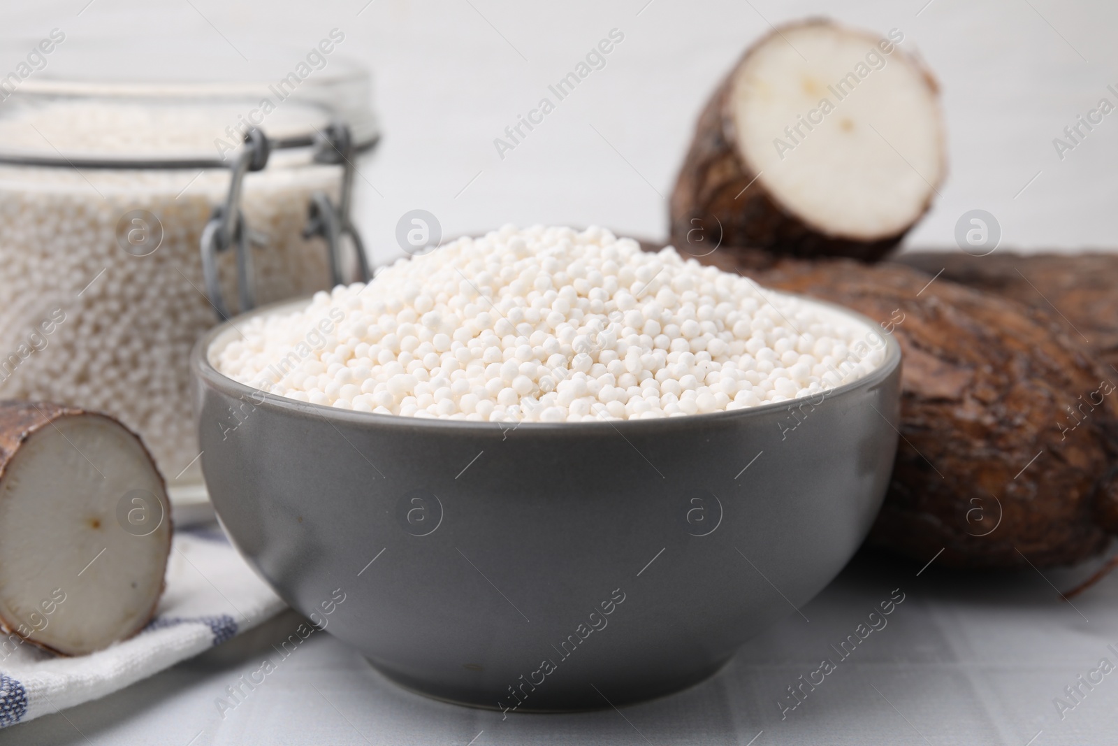 Photo of Tapioca pearls in bowl and cassava roots on white tiled table, closeup
