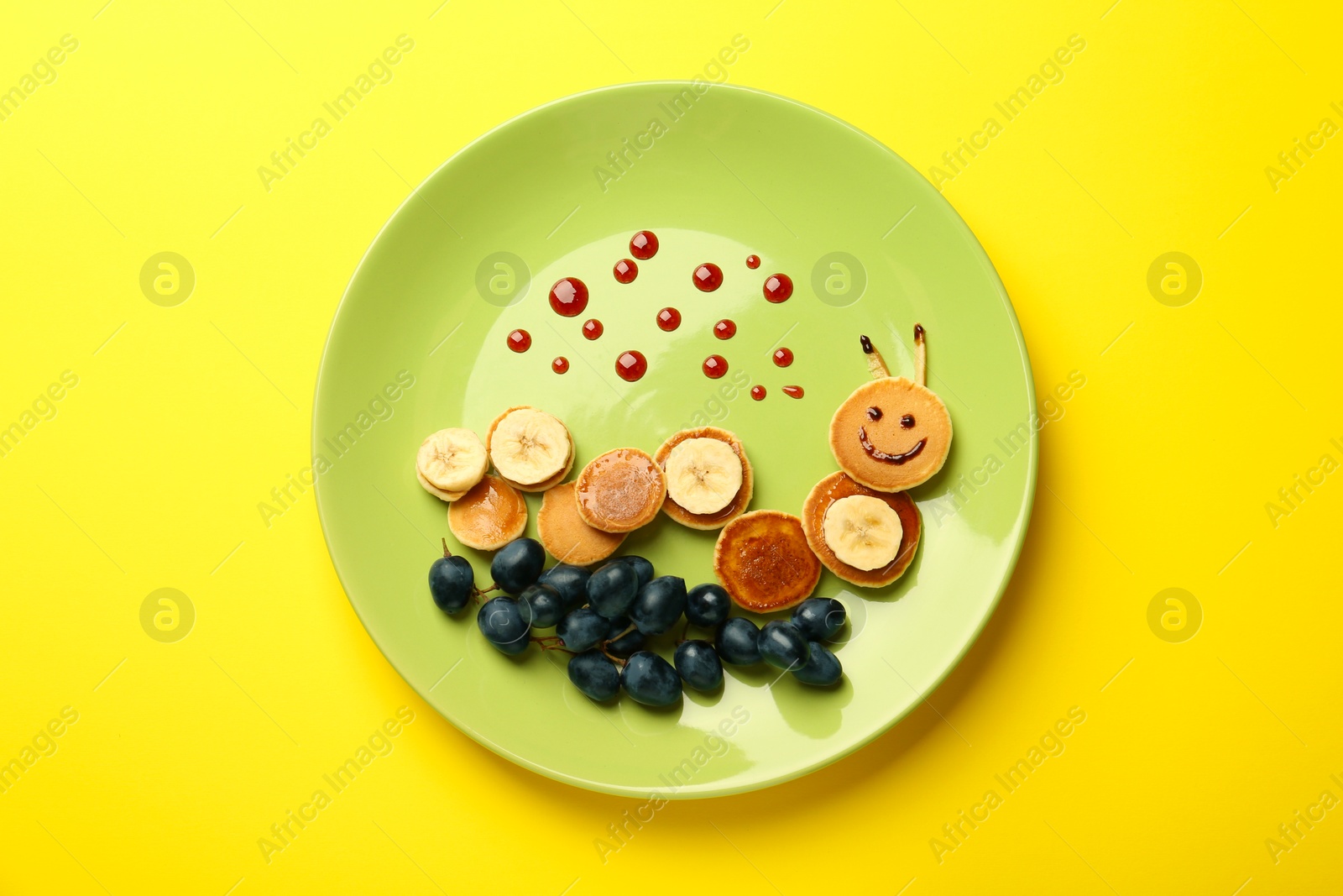 Photo of Creative serving for kids. Plate with cute caterpillar made of pancakes, grapes and banana on yellow background, top view