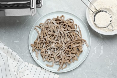 Photo of Uncooked homemade soba (buckwheat noodles), flour, napkin and pasta machine on grey table, flat lay