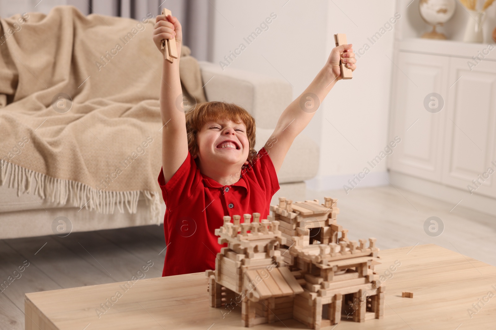 Photo of Emotional boy playing with wooden castle at table in room. Child's toy