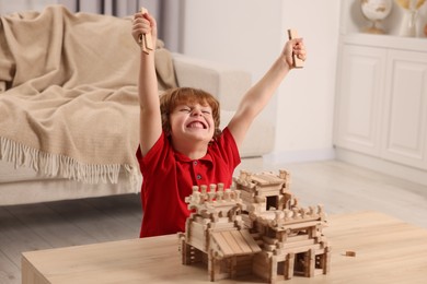 Photo of Emotional boy playing with wooden castle at table in room. Child's toy