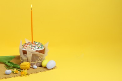 Traditional Easter cake with sprinkles, candle, decorated eggs and flowers on yellow background. Space for text