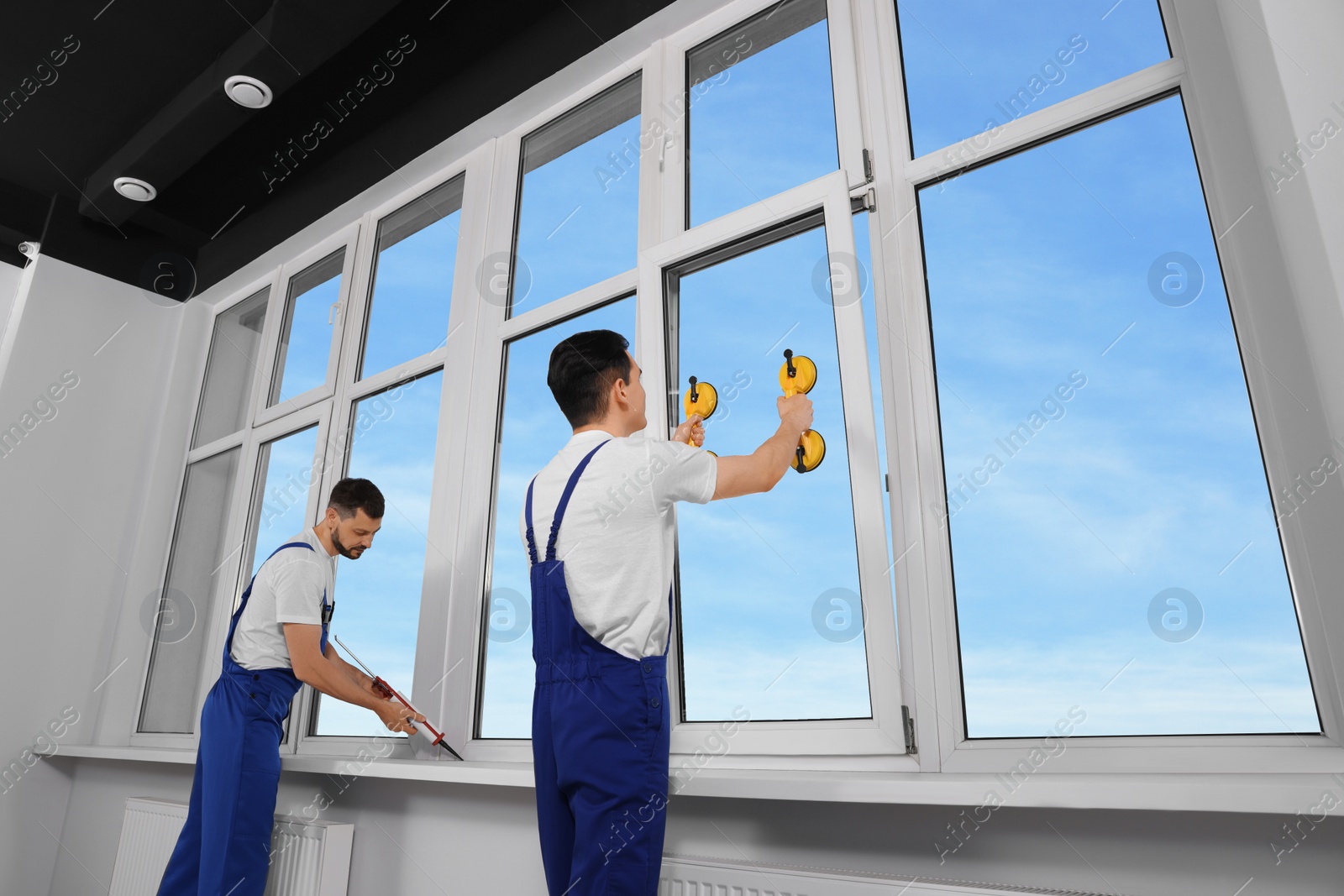 Photo of Workers with tube of sealant and suction lifters installing plastic windows indoors