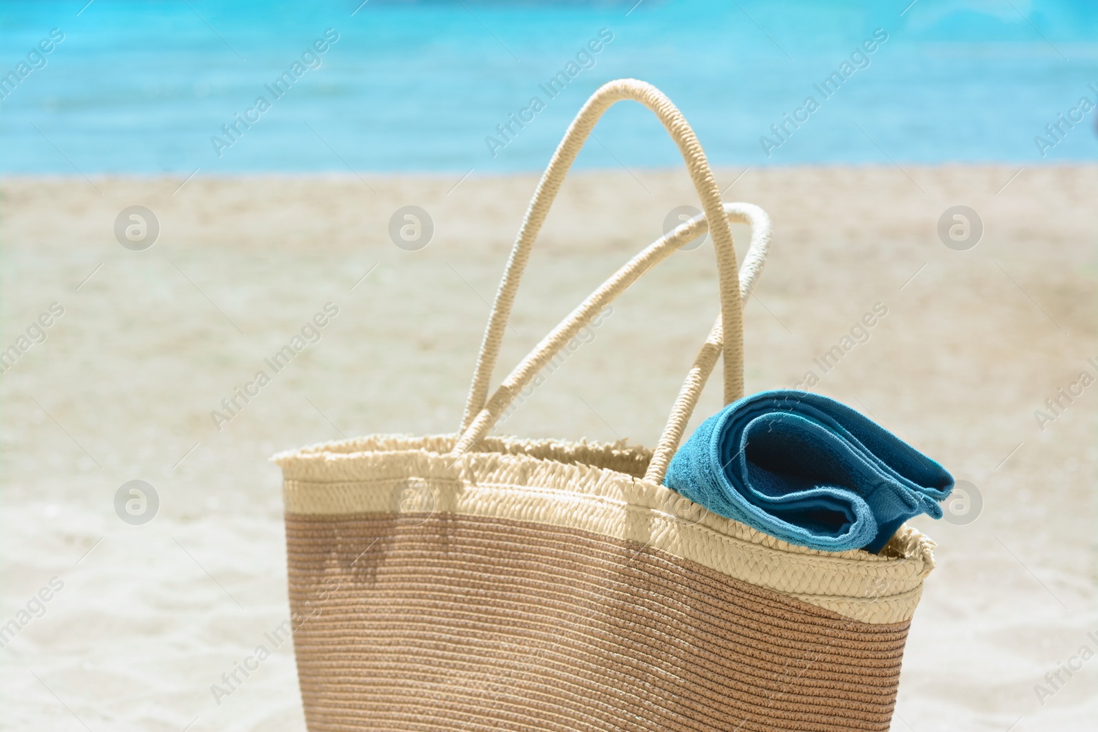 Photo of Soft blue towel in beach bag on sand near sea, closeup