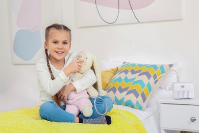 Photo of Little girl with toy bunny and nebulizer for inhalation on bed at home