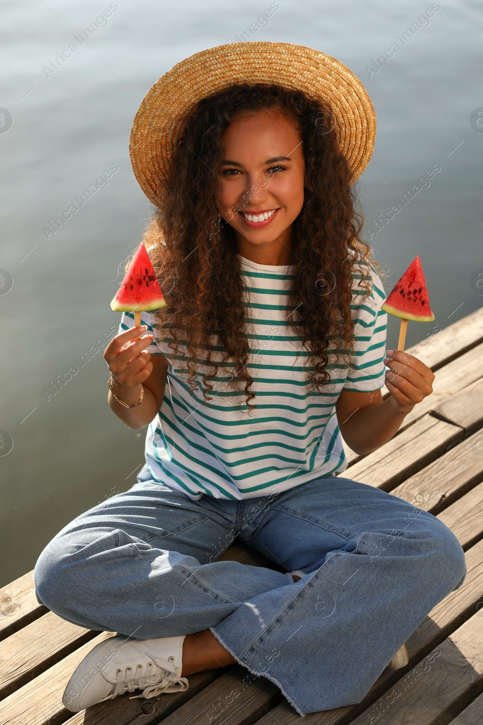 Photo of Beautiful young African American woman with pieces of watermelon on wooden pier near river