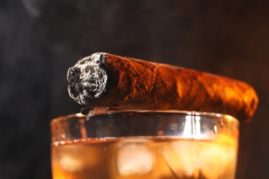 Photo of Glass of whiskey with ice cubes and smoldering cigar against black background, closeup