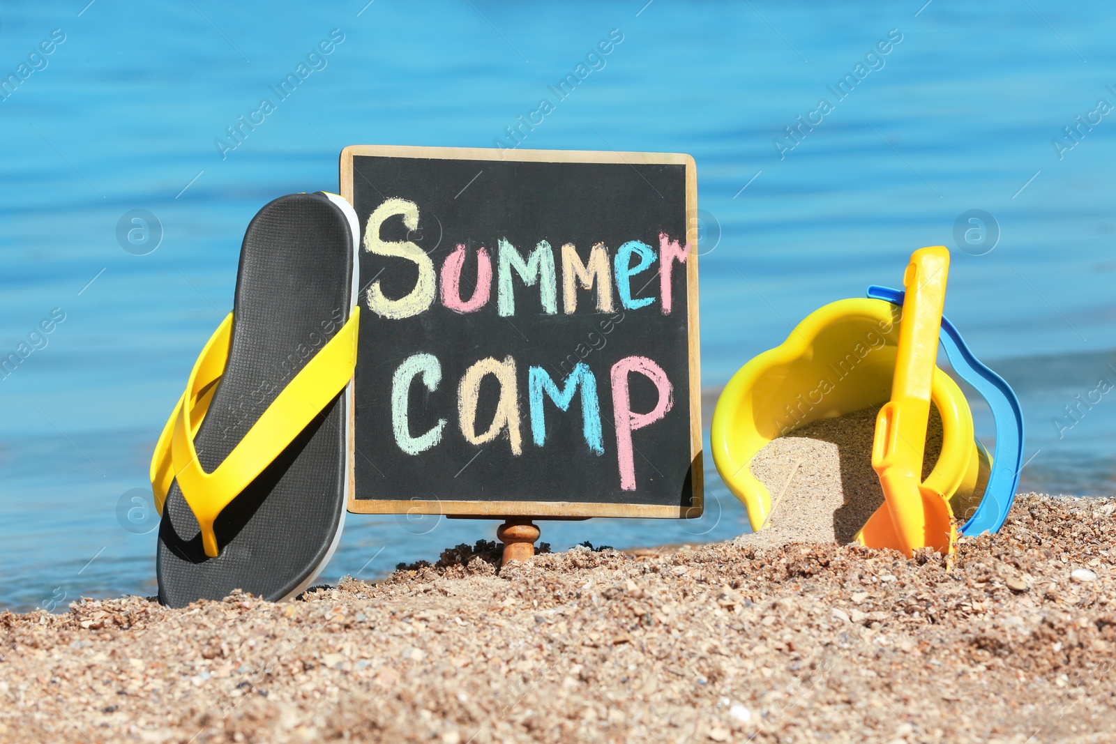 Photo of Little blackboard with text SUMMER CAMP, toys and flip flops on sand near sea