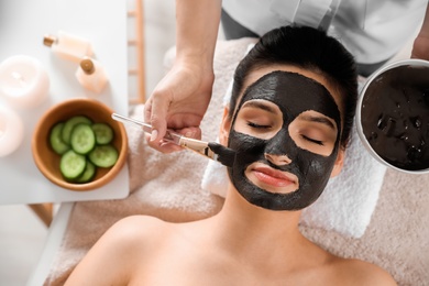 Photo of Cosmetologist applying black mask onto woman's face in spa salon, top view