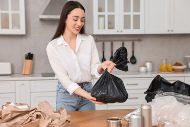 Photo of Garbage sorting. Smiling woman with plastic bag at table in kitchen