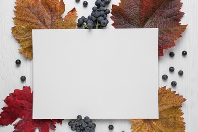 Flat lay composition with blank card, autumn leaves and grapes on white wooden table. Space for text