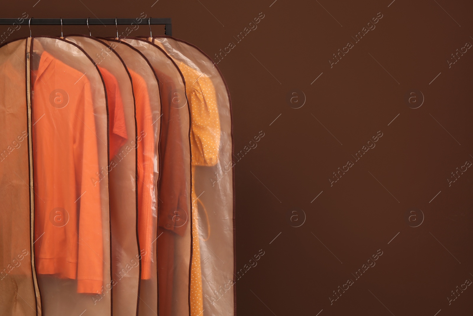 Photo of Garment bags with clothes on rack against brown background. Space for text