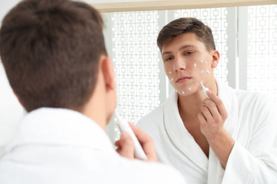 Teen guy with acne problem applying cream near mirror in bathroom