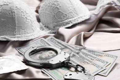 Photo of Prostitution concept. Handcuffs with dollar banknotes, condom and bra on wooden table, closeup