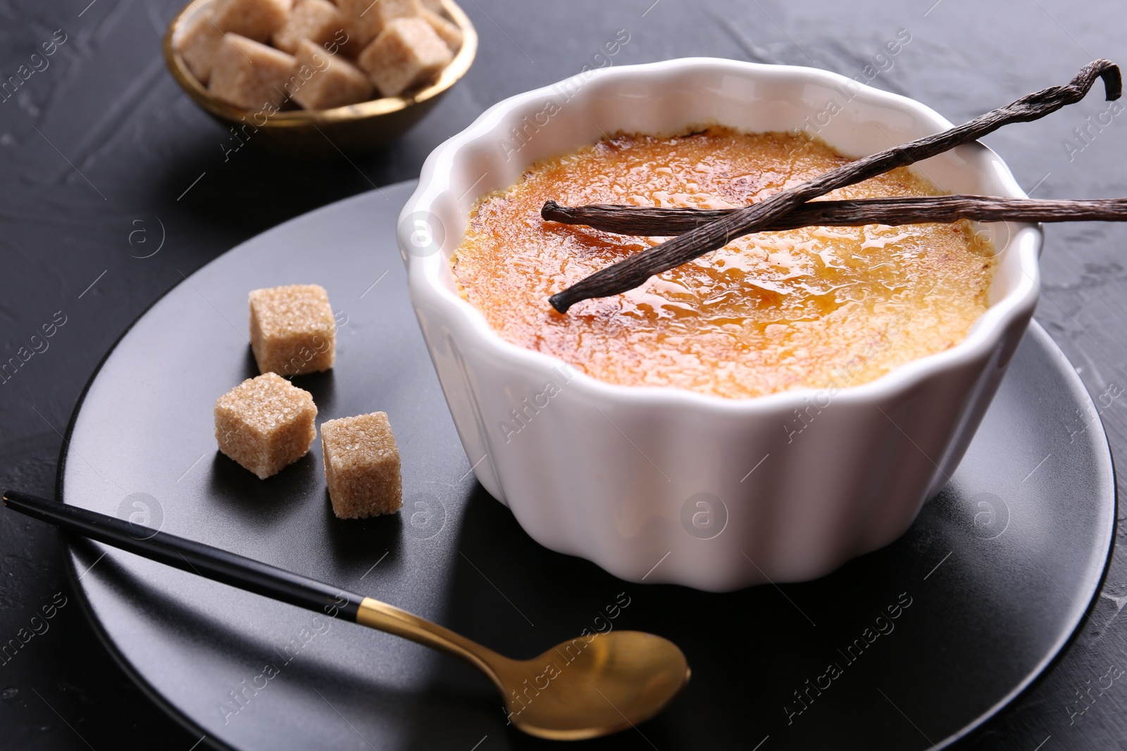 Photo of Delicious creme brulee in bowl, vanilla pods, sugar cubes spoon on dark gray table, closeup
