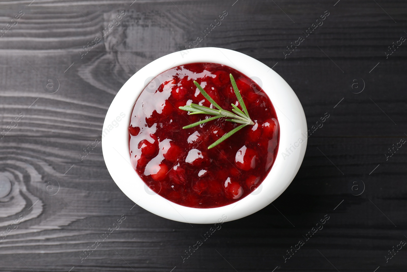 Photo of Fresh cranberry sauce in bowl and rosemary on black wooden table, top view