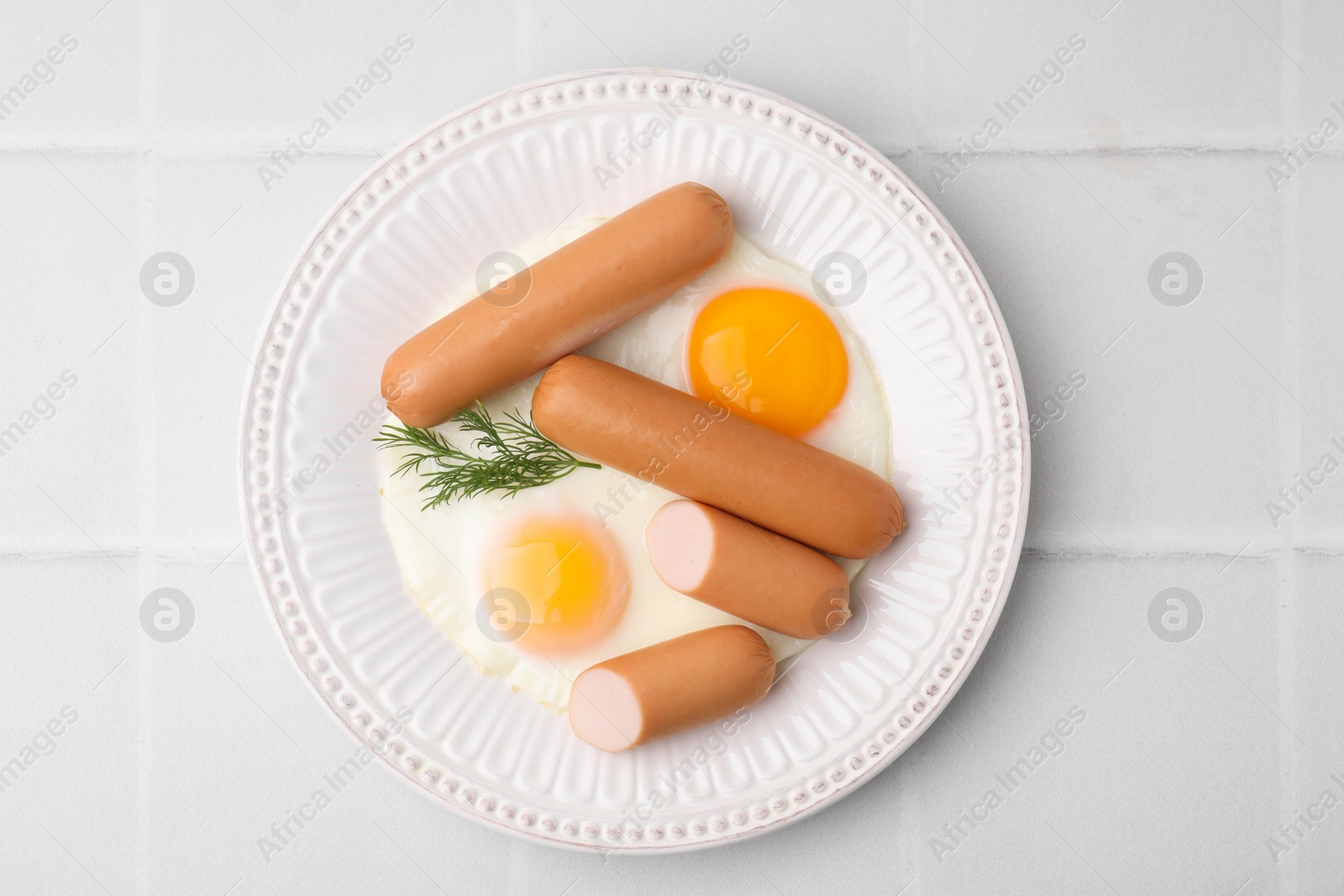 Photo of Delicious boiled sausages, fried eggs and dill on white tiled table, top view