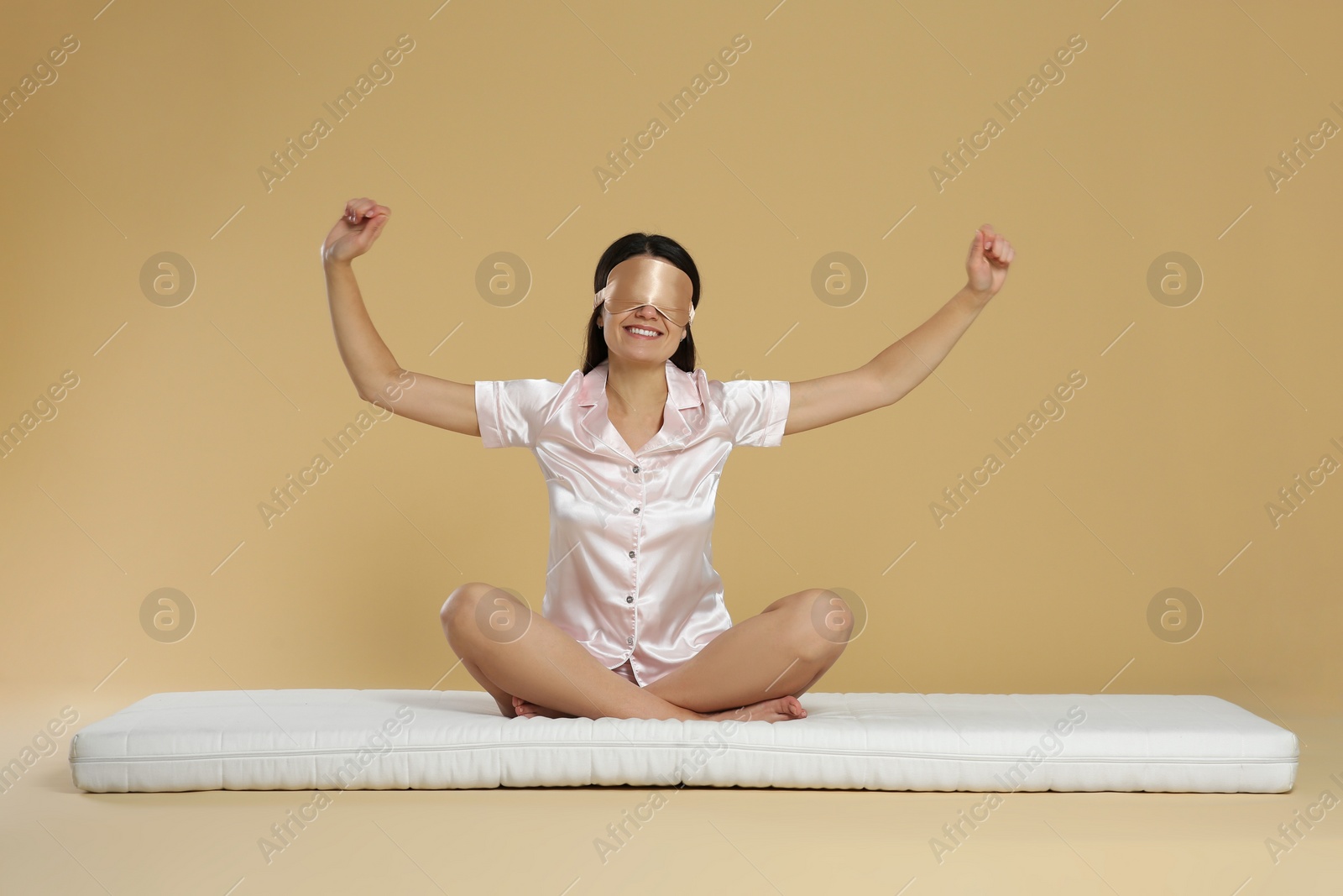 Photo of Woman with sleep mask stretching on soft mattress against beige background