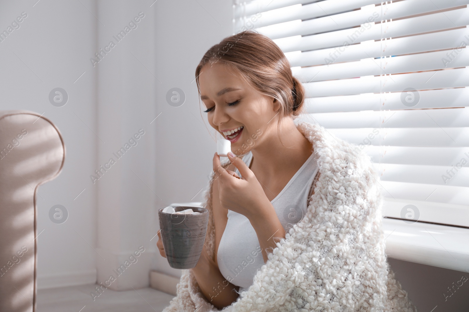 Photo of Beautiful young woman with cup of hot drink and marshmallow near window at home. Winter atmosphere