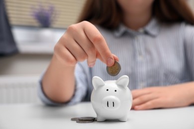 Woman putting coin into ceramic piggy bank at white wooden table indoors, closeup. Financial savings
