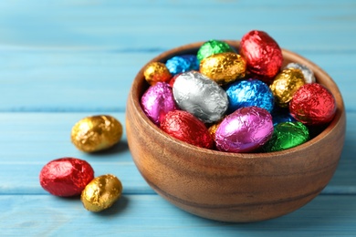 Photo of Bowl with chocolate eggs wrapped in colorful foil on light blue wooden table, closeup