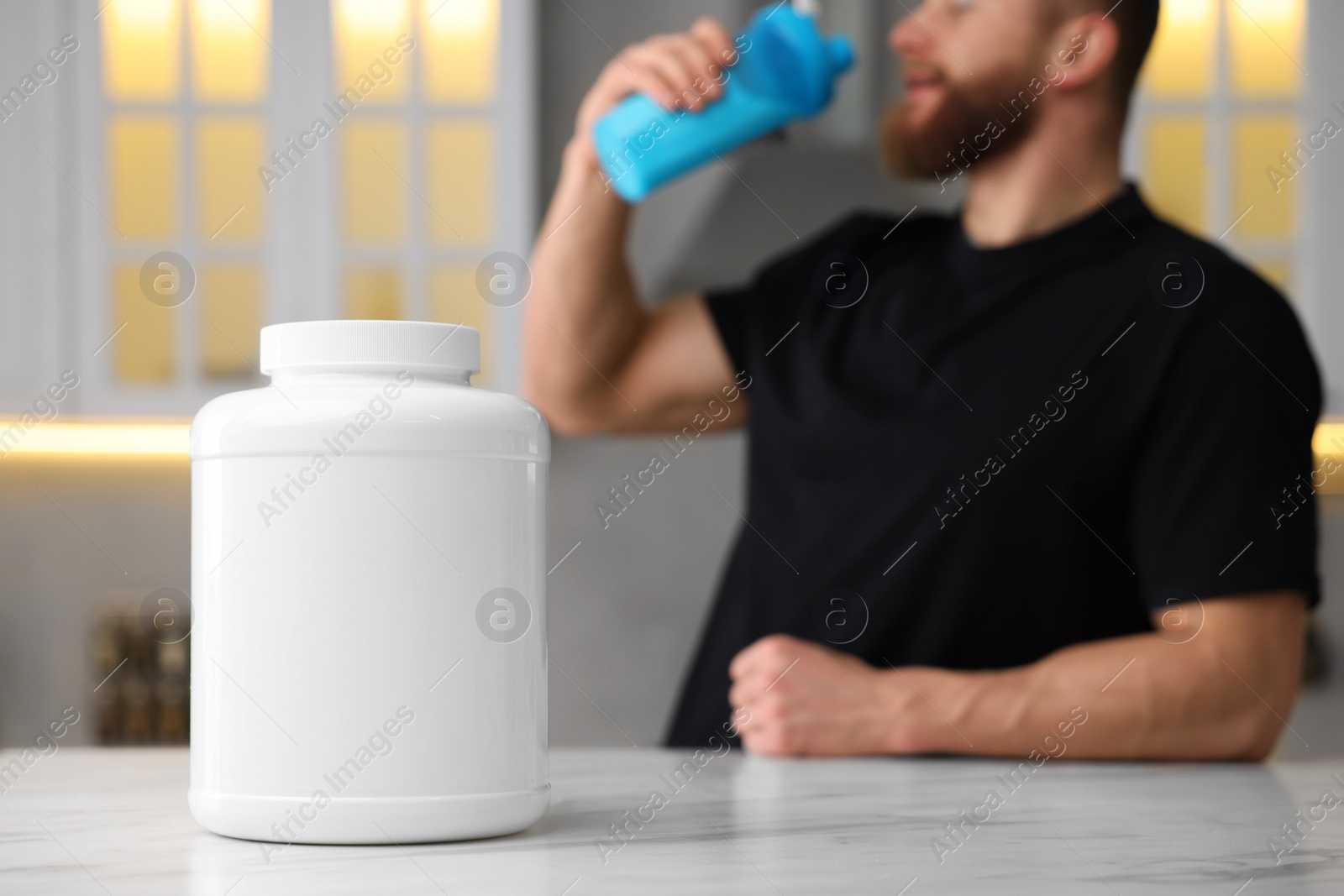 Photo of Young man with shaker of protein at white marble table in kitchen, focus on jar of powder