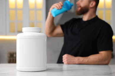 Young man with shaker of protein at white marble table in kitchen, focus on jar of powder