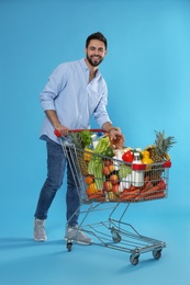 Photo of Young man with shopping cart full of groceries on light blue background