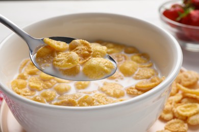 Photo of Spoon of delicious crispy corn flakes with milk above bowl on white table, closeup