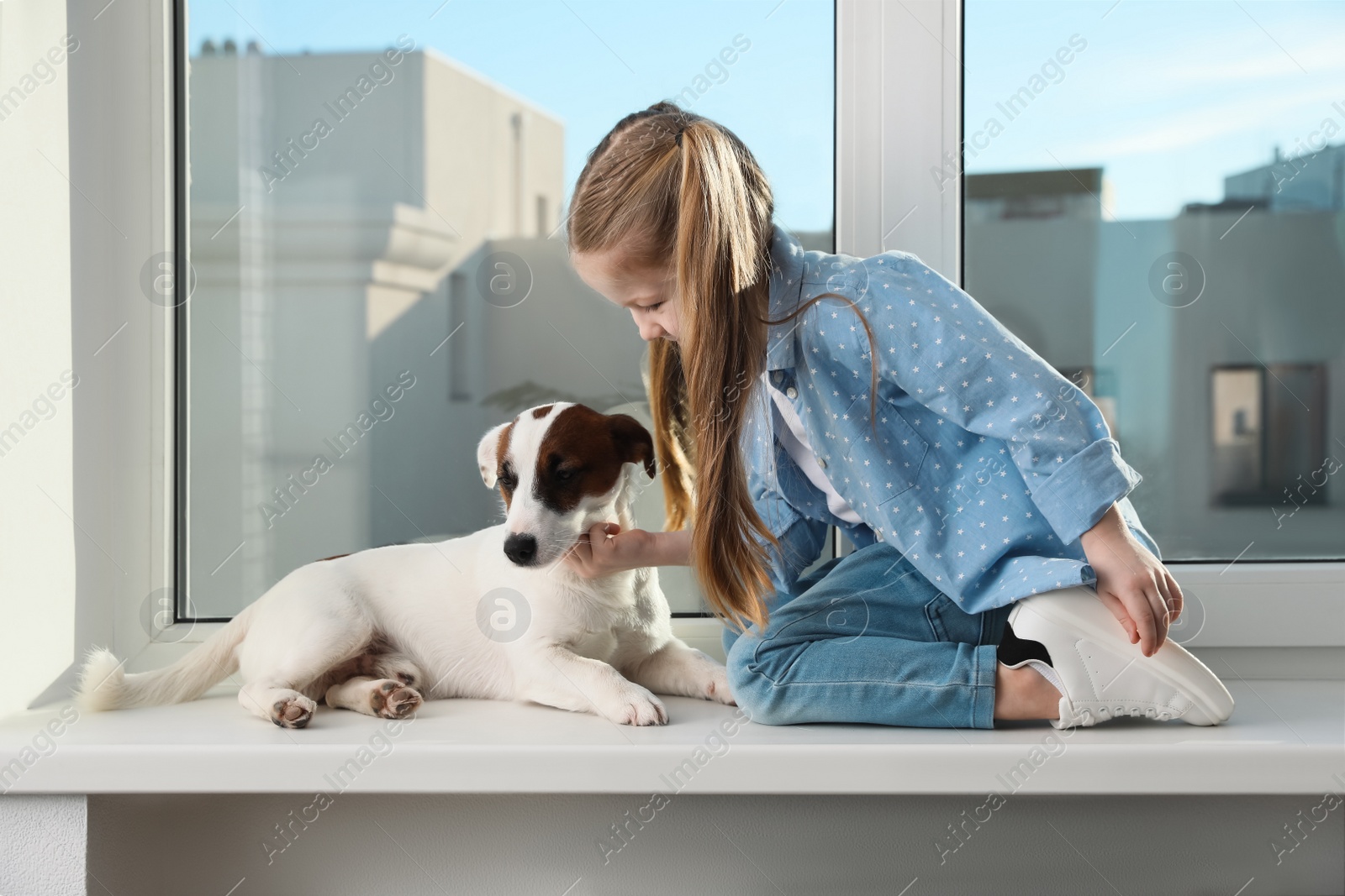 Photo of Cute little girl with her dog on window sill indoors. Childhood pet