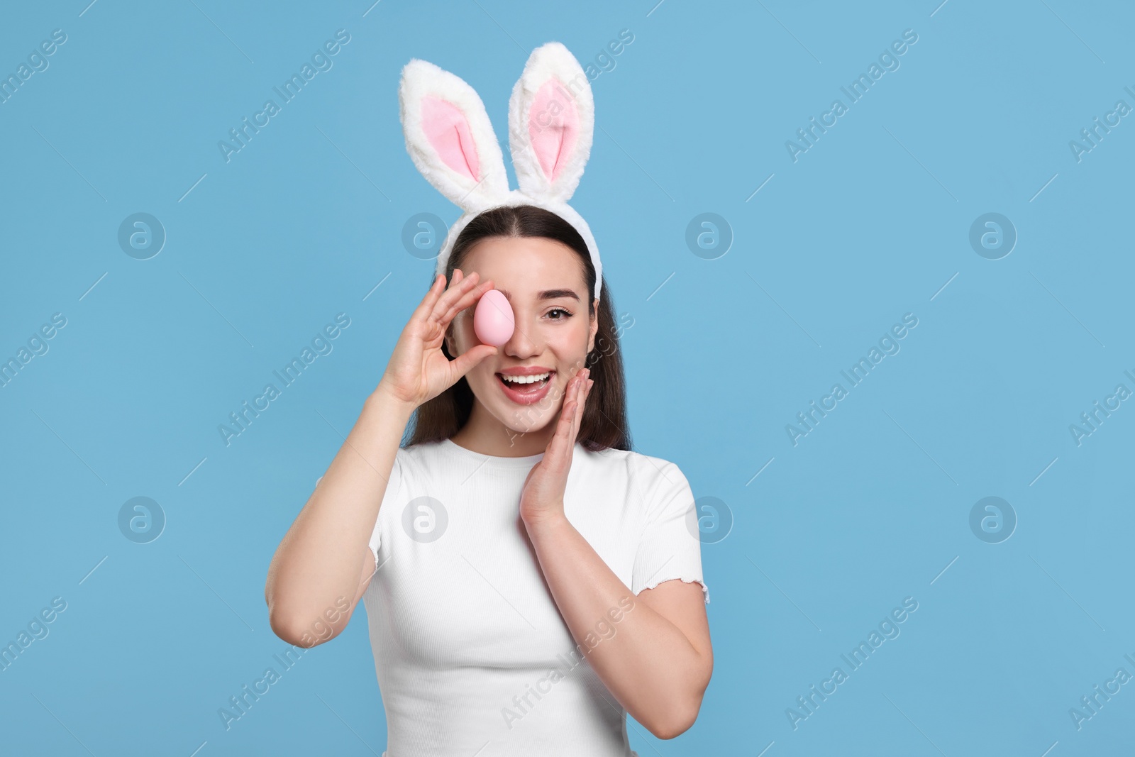 Photo of Happy woman in bunny ears headband holding painted Easter egg on turquoise background