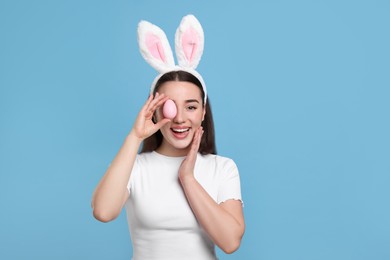 Photo of Happy woman in bunny ears headband holding painted Easter egg on turquoise background