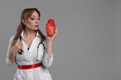 Photo of Woman in scary nurse costume with heart model on light grey background, space for text. Halloween celebration