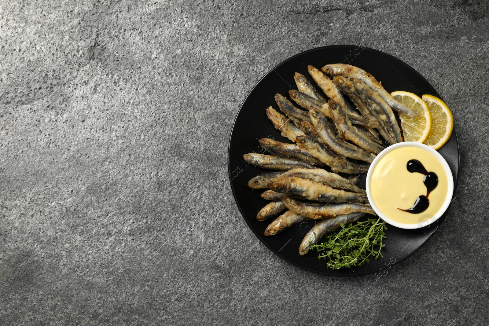 Photo of Plate with delicious fried anchovies, lemon, microgreens and sauce on grey table, top view. Space for text