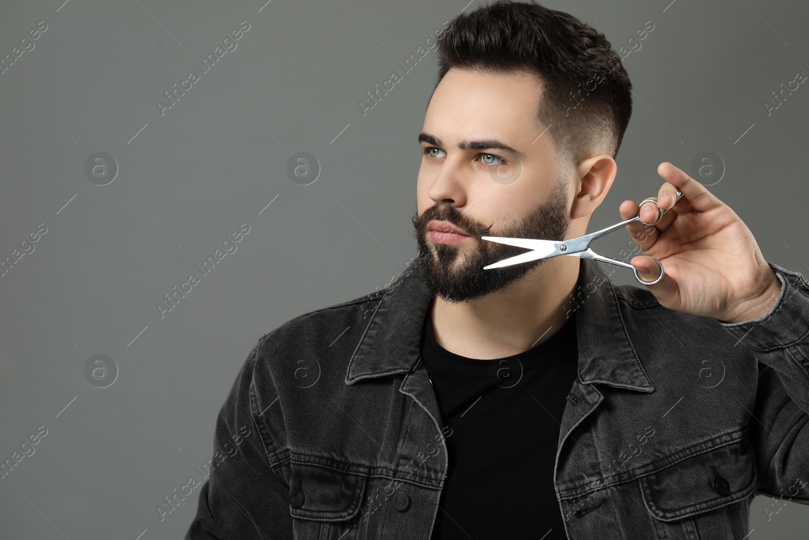 Photo of Handsome young man trimming beard with scissors on grey background. Space for text