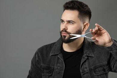 Photo of Handsome young man trimming beard with scissors on grey background. Space for text