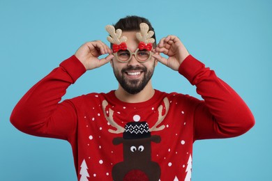 Happy young man in Christmas sweater and funny glasses on light blue background