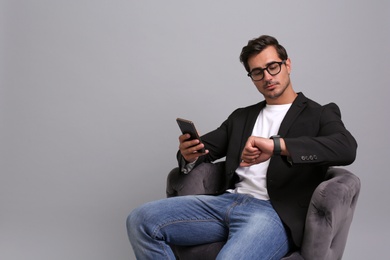 Handsome young businessman with smartphone checking time while sitting in armchair on grey background. Space for text