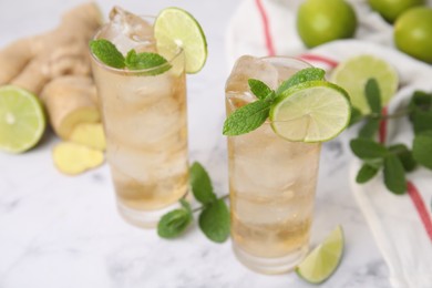 Glasses of tasty ginger ale with ice cubes and ingredients on white marble table, closeup