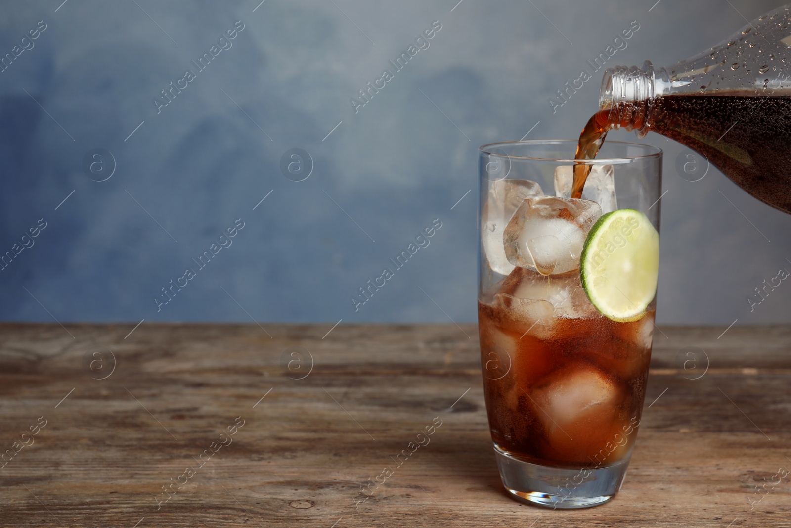 Photo of Pouring refreshing soda drink into glass with ice cubes and lime on wooden table against blue background. Space for text