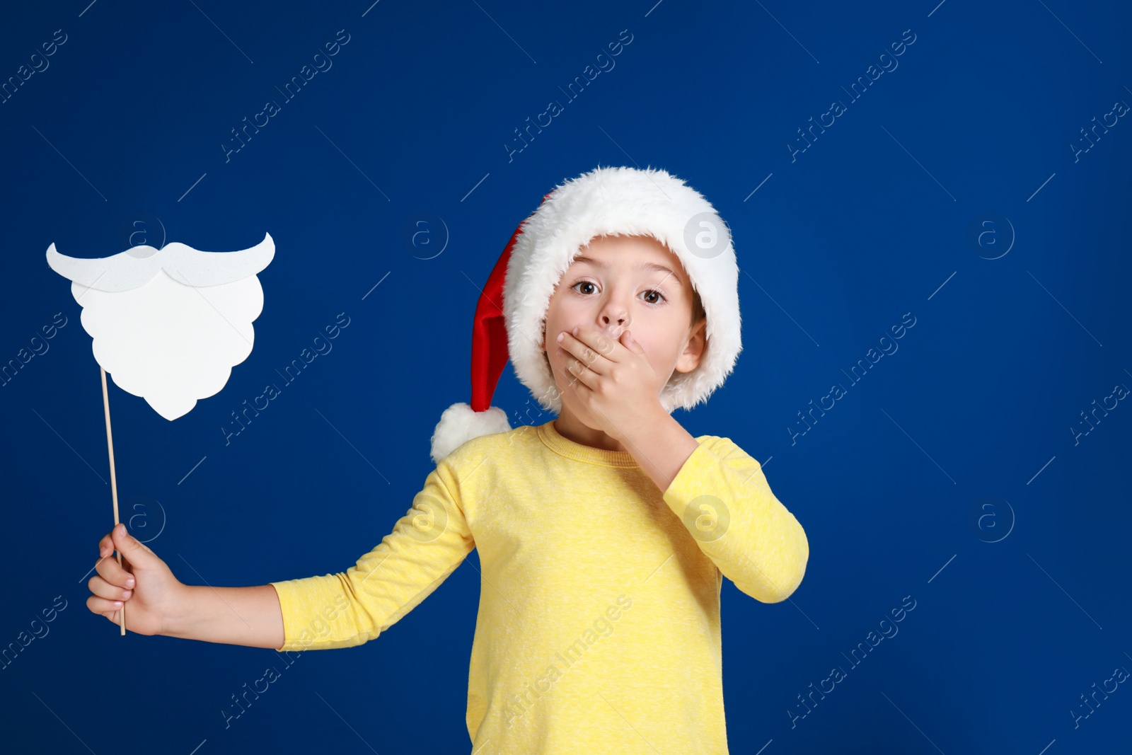 Image of Cute little boy with Santa hat and white beard prop on blue background. Christmas celebration