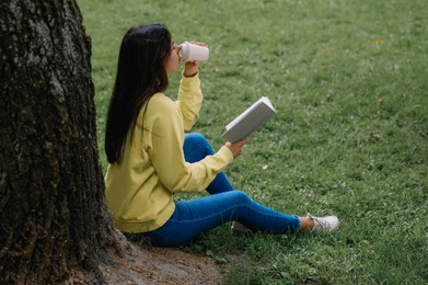 Photo of Young woman with cup of coffee reading book near tree in park