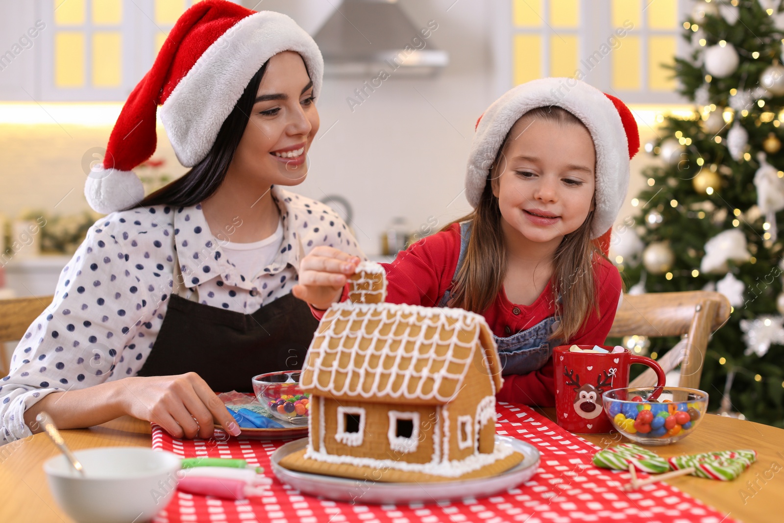 Photo of Mother and daughter decorating gingerbread house at table indoors